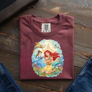 May include: Burgundy Comfort Colors t-shirt featuring a colorful illustration of two princesses, a deer, and a castle. The shirt has a crew neck and the Comfort Colors logo. The shirt is folded on a wooden surface.