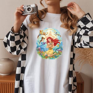 May include: White t-shirt featuring a colorful illustration of Disney princesses Ariel and Belle. Ariel, with red hair, is holding a small animal, while Belle reads a book. The design includes a castle, flowers, and woodland creatures.