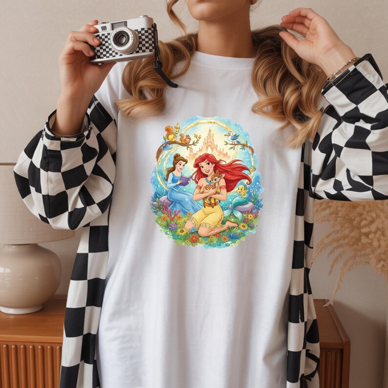 May include: White t-shirt featuring a colorful illustration of Disney princesses Ariel and Belle. Ariel, with red hair, is holding a small animal, while Belle reads a book. The design includes a castle, flowers, and woodland creatures.