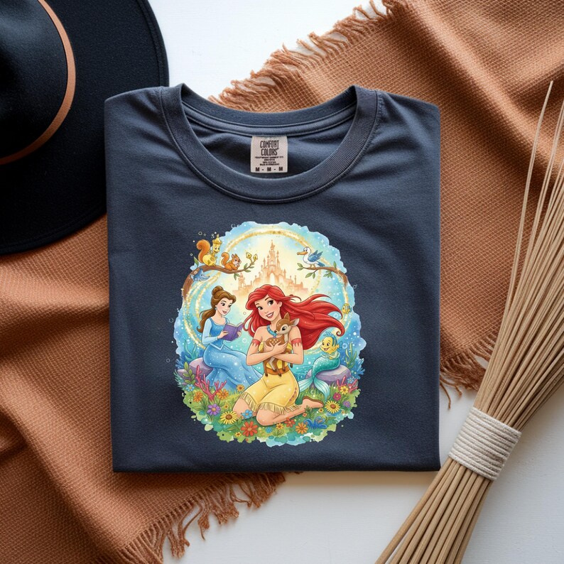 May include: Dark blue t-shirt featuring a colorful illustration of two princesses, a castle, and woodland creatures. The graphic is surrounded by a floral border. The shirt has a crew neck and short sleeves.