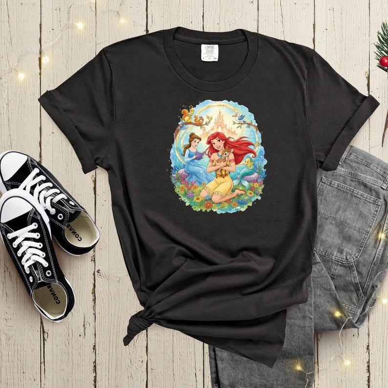 May include: Black t-shirt featuring a colorful graphic of two princesses in a floral setting with a castle in the background. The princesses are Ariel and Belle. The shirt is paired with black sneakers and gray jeans.