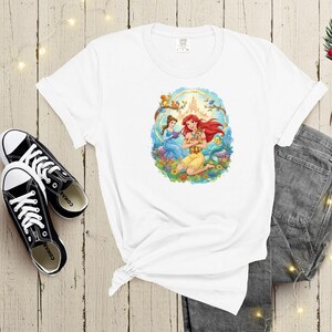 May include: White t-shirt featuring a colorful graphic of two princesses, one with red hair and the other with brown hair, surrounded by flowers and a castle. The shirt is paired with black sneakers and gray jeans.
