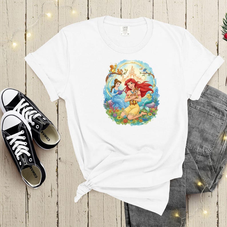May include: White t-shirt featuring a colorful graphic of two princesses, one with red hair and the other with brown hair, surrounded by flowers and a castle. The shirt is paired with black sneakers and gray jeans.