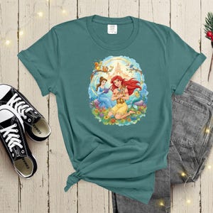 May include: Teal-colored t-shirt featuring a colorful illustration of two princesses, a fawn, and a mermaid. The graphic is surrounded by flowers, a castle, and woodland creatures. The shirt is paired with black sneakers and denim jeans.