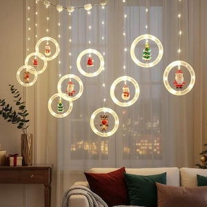 May include: A set of hanging LED string lights featuring multiple illuminated circular rings. Each ring contains a small Christmas-themed figurine, including Santa Claus, a snowman, a reindeer, and a miniature Christmas tree, suspended by thin wires.