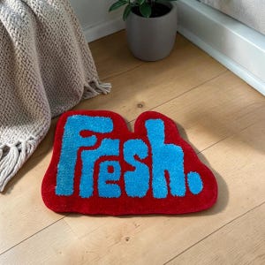 Peut inclure: Un tapis tufté de forme irrégulière et arrondie, présentant un fond rouge vif avec le mot "Fresh." écrit en grandes lettres bleues texturées. Le tapis possède une surface en poils doux et un design graphique audacieux et distinct.
