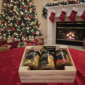 May include: A wooden crate holds three jars of honey with black lids and labels. The jars are labeled "Lemon", "Espresso", and "Creamed Cinnamon". The crate sits on a red tablecloth, with a Christmas tree and fireplace in the background.