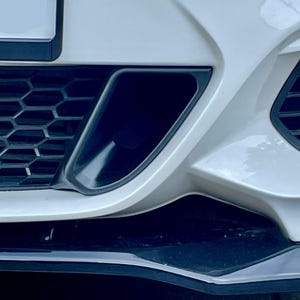 Może przedstawiać: Zbliżenie białego przedniego zderzaka samochodu, z czarnymi kratkami o strukturze plastra miodu i ciemnym wlotem powietrza. Zderzak ma elegancki, nowoczesny design z ostrymi kątami i czarną dolną wargą.