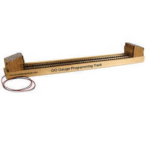 May include: A wooden OO Gauge Programming Track with two parallel tracks. The track has a light brown wooden base with the text "OO Gauge Programming Track" and a website address. Two brown and red wires are connected to the track.