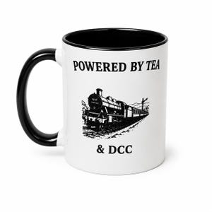 K&ouml;nnte beinhalten: Ein wei&szlig;er Keramikbecher mit schwarzem Henkel und schwarzem Innenrand. Auf der Vorderseite befindet sich eine schwarze Grafik einer Dampflokomotive auf Schienen mit dem Text POWERED BY TEA dar&uuml;ber und & DCC darunter in Serifenschrift.