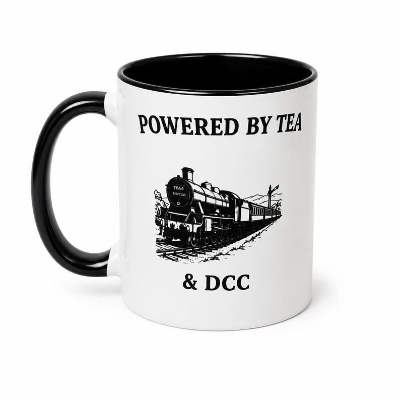 K&ouml;nnte beinhalten: Ein wei&szlig;er Keramikbecher mit schwarzem Henkel und schwarzem Innenrand. Auf der Vorderseite befindet sich eine schwarze Grafik einer Dampflokomotive auf Schienen mit dem Text POWERED BY TEA dar&uuml;ber und & DCC darunter in Serifenschrift.