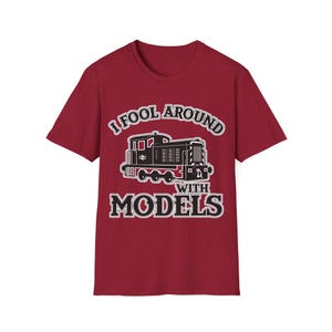 Könnte beinhalten: Ein dunkelrotes Kurzarm-T-Shirt mit einer schwarz-weißen Lokomotivgrafik. Der Text "I FOOL AROUND WITH MODELS" ist in einer fetten, weiß umrandeten Schriftart oberhalb und unterhalb der Zugillustration auf der Vorderseite aufgedruckt.