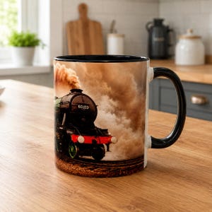 Może przedstawiać: Ceramiczny kubek z czarnym uchwytem i obrzeżem, ozdobiony fotograficznym nadrukiem czarnej lokomotywy parowej o numerze 60103. Pociąg znajduje się na torach i wypuszcza dużą chmurę brązowego dymu na tle w odcieniach sepii.