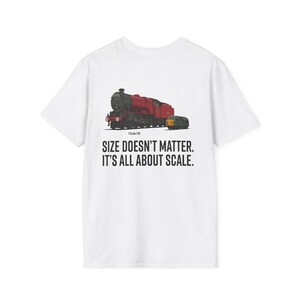 Può includere: T-shirt bianca con grafica di un treno a vapore rosso e nero e un vagone passeggeri giallo. Il testo "SIZE DOESN'T MATTER. IT'S ALL ABOUT SCALE." è stampato sotto. La maglietta è in materiale morbido.