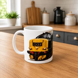 Peut inclure: Un mug en c&eacute;ramique blanche orn&eacute; d'une illustration d&eacute;taill&eacute;e d'une locomotive diesel jaune et noire. L'avant du train affiche le num&eacute;ro 037 dans un encadr&eacute; rectangulaire noir. Le motif pr&eacute;sente des d&eacute;tails d'usure r&eacute;alistes sur la carrosserie.