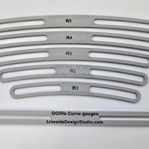 Pode incluir: Um conjunto de medidores de curva cinza, incluindo cinco medidores curvos rotulados de R1 a R5 e um medidor reto. Os medidores têm medidas em milímetros. O texto "OO/Ho Curve gauges" e "LinesideDesignStudio.com" é impresso no medidor reto.