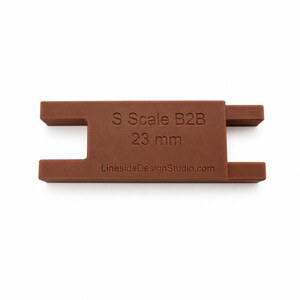 Peut inclure: Un outil de mesure S Scale B2B 23 mm marron avec un corps rectangulaire et deux d&eacute;coupes en forme de U sur les c&ocirc;t&eacute;s. Le texte S Scale B2B 23 mm et LinesideDesignStudio.com est grav&eacute; sur la face avant de cet outil de mod&eacute;lisme ferroviaire.