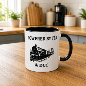 K&ouml;nnte beinhalten: Ein wei&szlig;er Keramikbecher mit schwarzem Innenraum und schwarzem Henkel. Auf der Vorderseite befindet sich eine schwarze grafische Abbildung einer Dampflokomotive auf Schienen, mit dem Text "POWERED BY TEA" dar&uuml;ber und "& DCC" darunter.