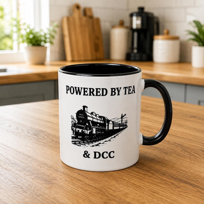 K&ouml;nnte beinhalten: Ein wei&szlig;er Keramikbecher mit schwarzem Innenraum und schwarzem Henkel. Auf der Vorderseite befindet sich eine schwarze grafische Abbildung einer Dampflokomotive auf Schienen, mit dem Text "POWERED BY TEA" dar&uuml;ber und "& DCC" darunter.
