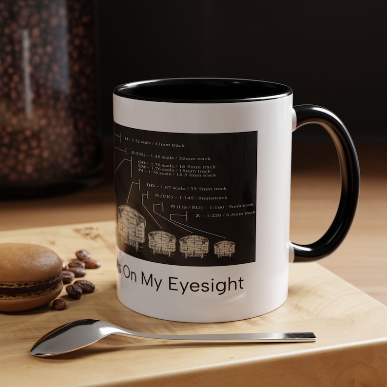 May include: A white ceramic mug with a black interior and handle. The exterior features a black rectangular graphic displaying various model train track scales and measurements, with the text "On My Eyesight" printed in black below the graphic.