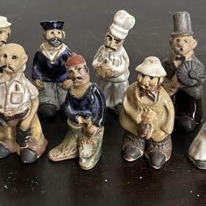 Op de afbeelding: Een verzameling kleine, handgeschilderde keramische beeldjes die verschillende beroepen en personages uitbeelden. De beeldjes zijn in verschillende kleuren, waaronder bruin, beige, blauw en rood. Elk beeldje is ongeveer 7,6 cm hoog.