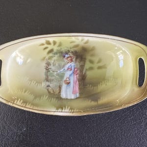 May include: Oval-shaped ceramic dish with handles, featuring a hand-painted scene of a woman in a blue dress and pink bow, carrying a basket in a pastoral setting. The dish has a gold rim and is approximately 8 inches long.