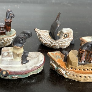 May include: Five miniature ceramic boats in various colors, including white, brown, and green. Each boat has unique details, such as smokestacks and decorative patterns. The boats are arranged on a dark surface.