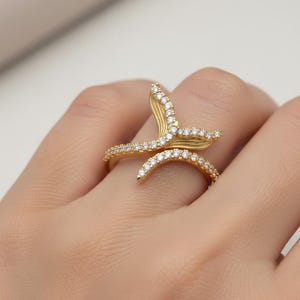 Op de afbeelding: Een goudkleurige ring met een uniek, vloeiend ontwerp. De ring is voorzien van een reeks kleine, heldere edelstenen die de rondingen van het ontwerp volgen. De vorm van de ring doet denken aan een gestileerde golf of een plantenrank.