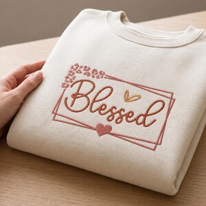 Könnte beinhalten: Ein cremefarbenes Sweatshirt mit Rundhalsausschnitt und einer Stickerei auf der Vorderseite. Die Stickerei zeigt das Wort Blessed in Schreibschrift, eingerahmt von einem rechteckigen Rahmen, verziert mit einem kleinen Herz und Leopardenmuster.