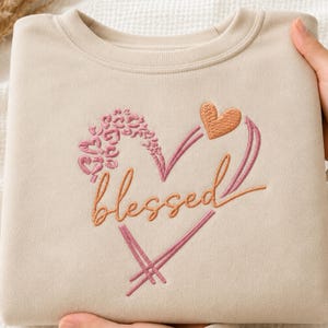 May include: A beige crewneck sweatshirt featuring an embroidered design of a heart outline in pink and orange thread. The word blessed is stitched in orange cursive script across the center, with a small orange heart and pink leopard print accents.
