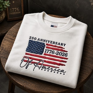 May include: A white crewneck sweatshirt featuring an embroidered design of a stylised American flag with the text 250 ANNIVERSARY 1776-2026 Of America. The embroidery uses navy blue and red thread, and the garment has a small flag tag inside the collar.