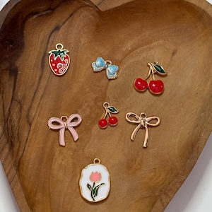 Puede incluir: Una bandeja de madera en forma de corazón contiene varios pequeños abalorios. Incluyen una fresa roja, un lazo azul, cerezas rojas, un lazo rosa, cerezas rojas y un lazo dorado. También hay un abalorio de tulipán blanco y dorado.