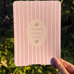 May include: A rectangular card with a light pink and white vertical striped pattern. In the centre, an oval frame with a scalloped edge features small pink floral accents and the text currently escaping reality in a simple font.
