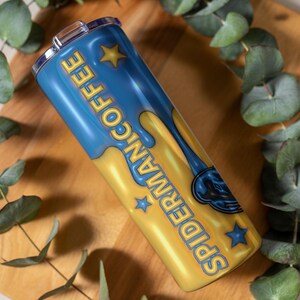 May include: A blue and yellow tumbler with the words "Spider-Man Coffee" in yellow lettering. The tumbler has a silver lid and is decorated with star shapes. The tumbler is resting on a wooden surface with green leaves.