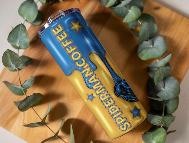 May include: A blue and yellow tumbler with the words "Spider-Man Coffee" in yellow lettering. The tumbler has a silver lid and is decorated with star shapes. The tumbler is resting on a wooden surface with green leaves.