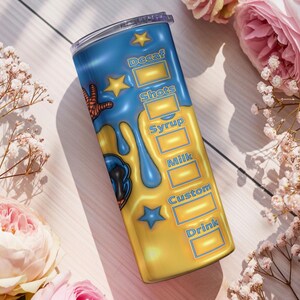 May include: A blue and yellow insulated tumbler with a cartoon design and the words "Decaf", "Shots", "Syrup", "Milk", "Custom", and "Drink". The tumbler has a clear lid and is decorated with stars and a cartoon character. The background includes flowers.