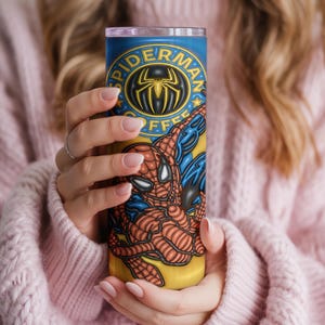 May include: A tall, cylindrical tumbler with a Spider-Man design. The tumbler has a blue background with the words "Spider-Man Coffee" and a Spider-Man logo. The superhero is shown in his classic red and blue suit.