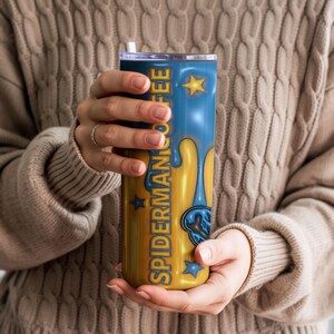 May include: A blue and gold tumbler with the text "SPIDERMAN COFFEE" and a dripping design. The tumbler has a straw and is held by a person wearing a beige jumper. The design includes stars and a Spider-Man graphic.