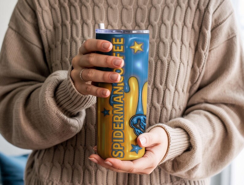 May include: A blue and gold tumbler with the text "SPIDERMAN COFFEE" and a dripping design. The tumbler has a straw and is held by a person wearing a beige jumper. The design includes stars and a Spider-Man graphic.