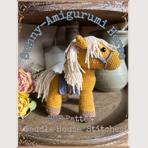 May include: A mustard yellow crocheted amigurumi horse with a cream-colored yarn mane and tail, wearing a blue saddle and bridle. The text Sunny-Amigurumi Horse, PDF Pattern, and Saddle House Stitches is printed in blue over the image.