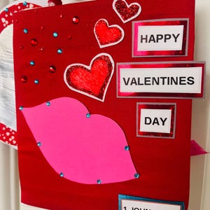 May include: A red Valentine's Day gift bag with a pink lip design and glittery hearts. The bag features the words "HAPPY VALENTINES DAY" and the Bible verse 1 John 4: 7-8.