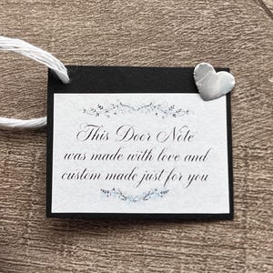 May include: A rectangular black tag with a white center panel featuring the text "This Door Note was made with love and custom made just for you" in black script, accented by floral flourishes and a small silver-colored metal heart charm.