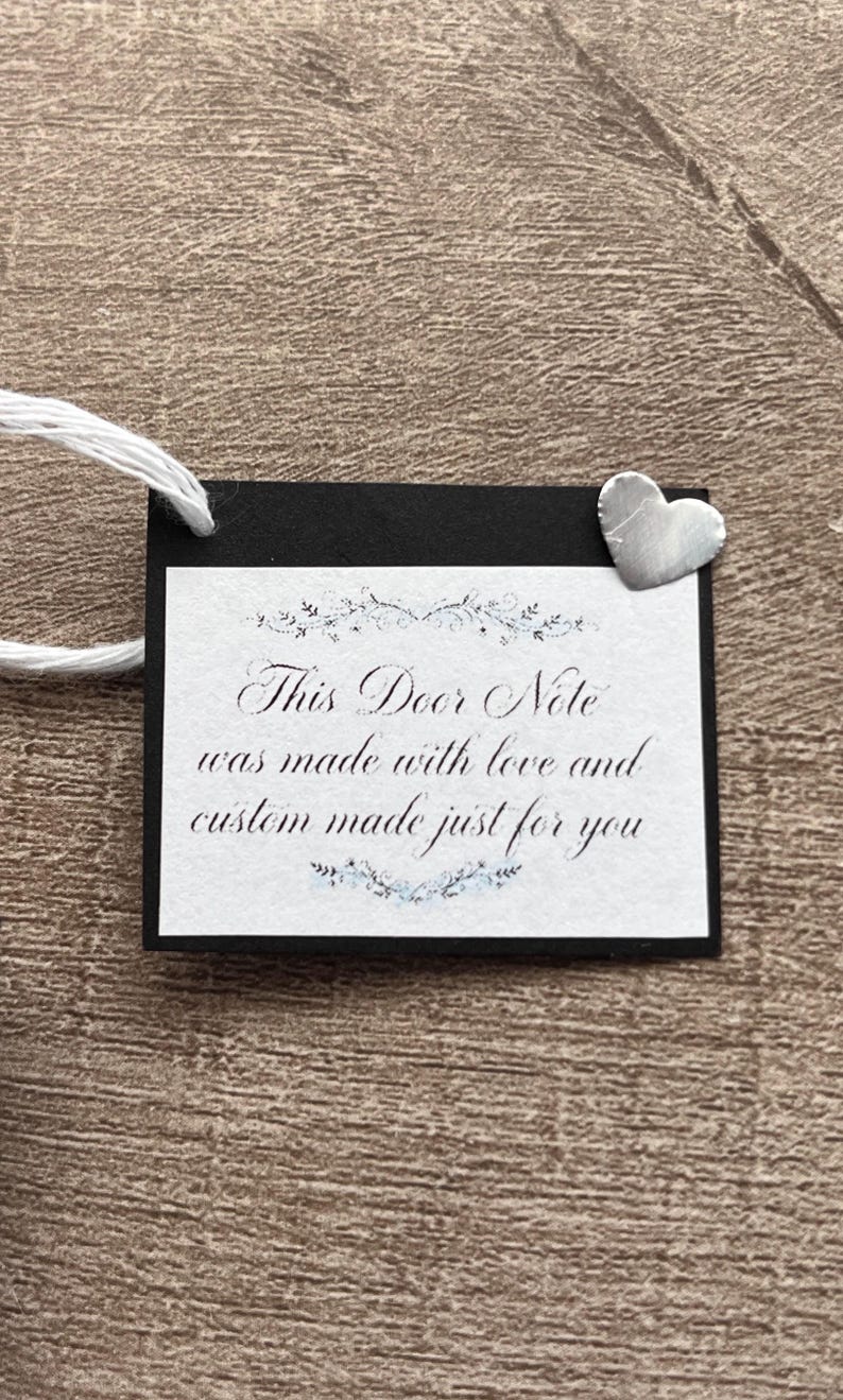 May include: A rectangular black tag with a white center panel featuring the text "This Door Note was made with love and custom made just for you" in black script, accented by floral flourishes and a small silver-colored metal heart charm.