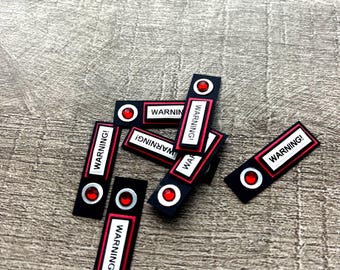 May include: A collection of rectangular black labels featuring the text WARNING! in black letters on a white background, bordered by a red frame. Each label includes a circular red rhinestone accent at one end, creating a decorative warning sign.