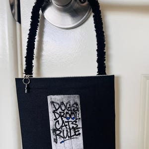 Puede incluir: Colgador de puerta negro con el texto "DOGS DROOL CATS RULE" en estilo grafiti. El colgador está sujeto a una manija de puerta con un limpiapipas negro y difuso. También son visibles tres imanes circulares rojos y una etiqueta que dice "I PREFER DROOL".
