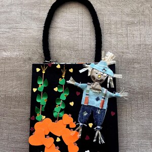 May include: A black rectangular bag with a black fuzzy handle featuring a scarecrow figure, orange paper hearts, and green paper vines. The bag includes a small label at the bottom with the text MATHEW 9:35-38 printed in black on a white background.