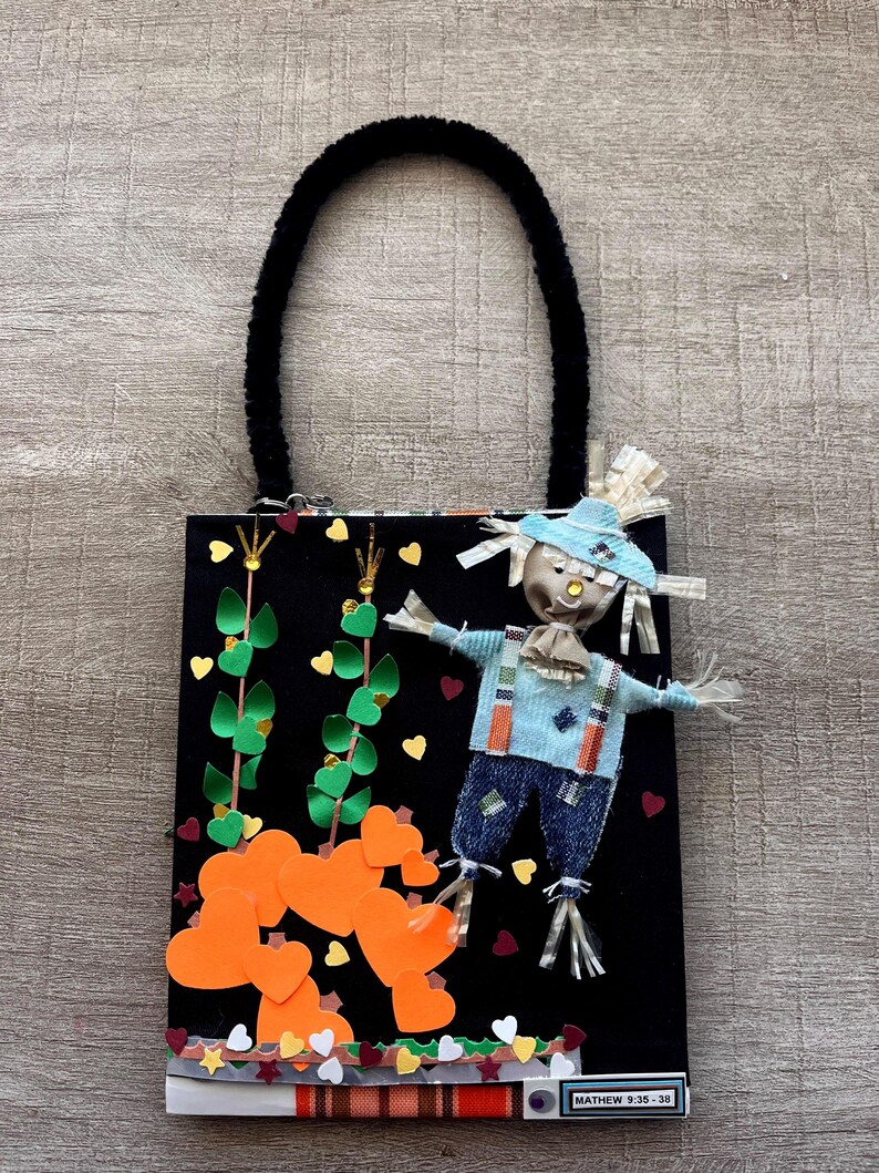 May include: A black rectangular bag with a black fuzzy handle featuring a scarecrow figure, orange paper hearts, and green paper vines. The bag includes a small label at the bottom with the text MATHEW 9:35-38 printed in black on a white background.