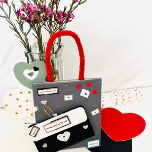 Può includere: Una borsa regalo grigia fatta a mano con un manico rosso, decorata con piccoli cuori rossi e note. È allegata una busta bianca e nera con la scritta "TO THE LOVE OF MY LIFE". Un vaso di fiori rosa è sullo sfondo.