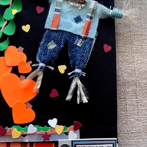 May include: A decorative wall hanging on a black background featuring a scarecrow figure with a light blue shirt, denim pants, and straw accents. The scene includes orange heart-shaped cutouts, green leaves, and a label reading MATHEW 9:35-38.
