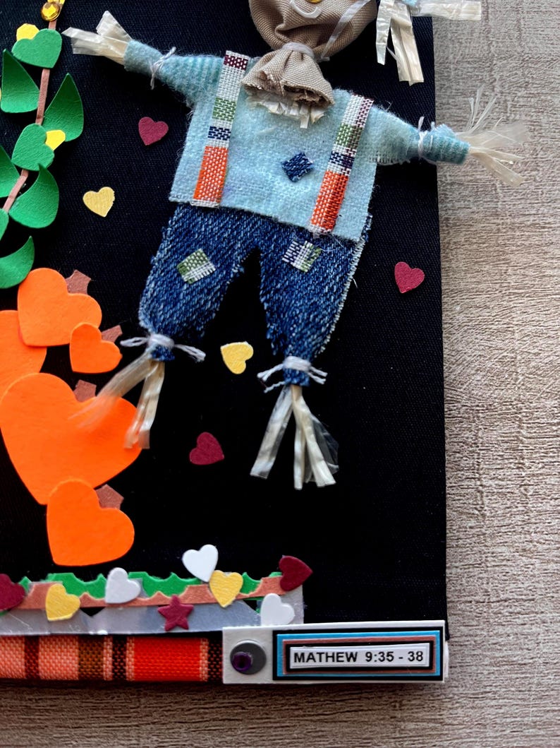 May include: A decorative wall hanging on a black background featuring a scarecrow figure with a light blue shirt, denim pants, and straw accents. The scene includes orange heart-shaped cutouts, green leaves, and a label reading MATHEW 9:35-38.